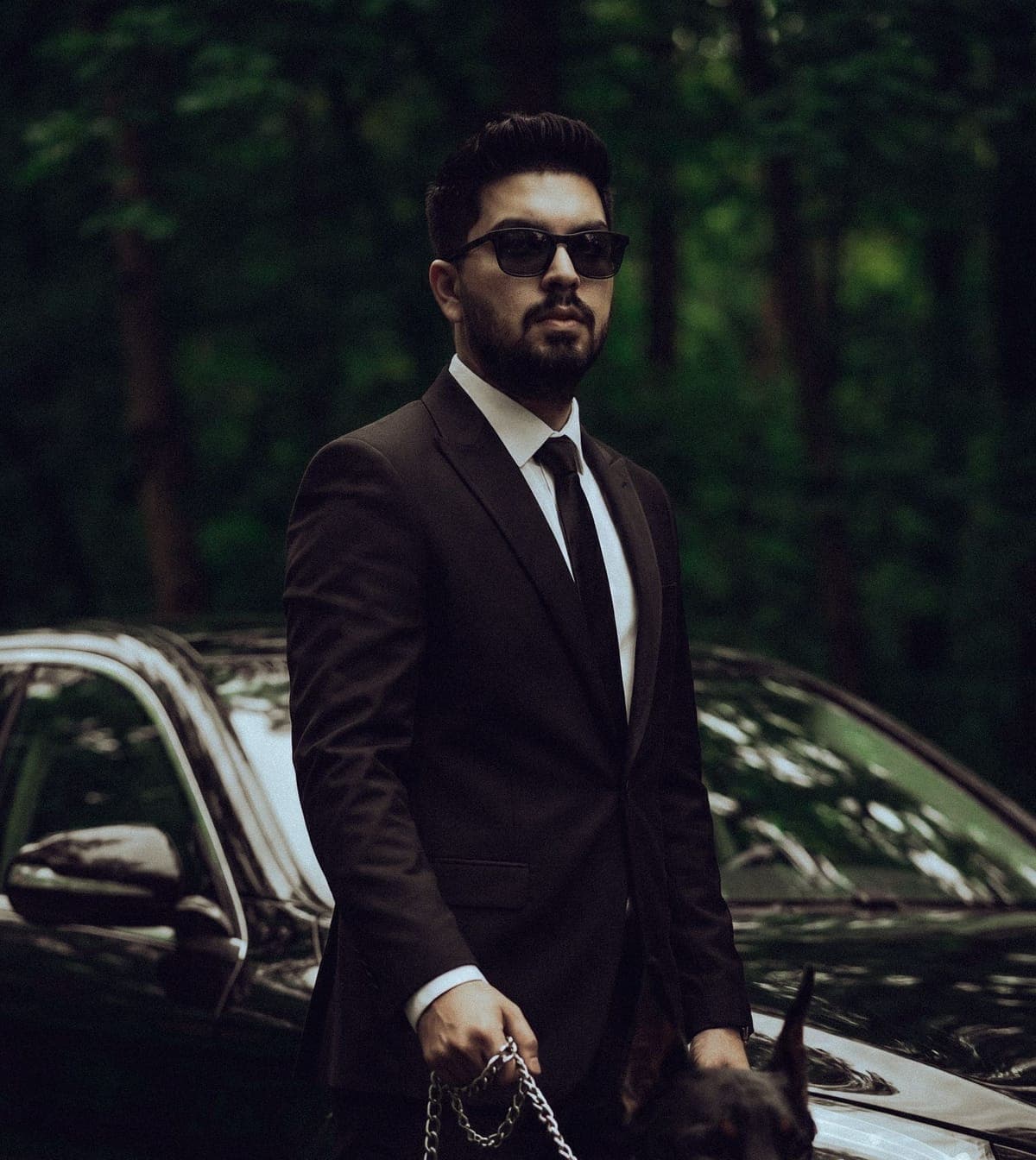 A bodyguard in a dark suit and sunglasses standing beside a black car