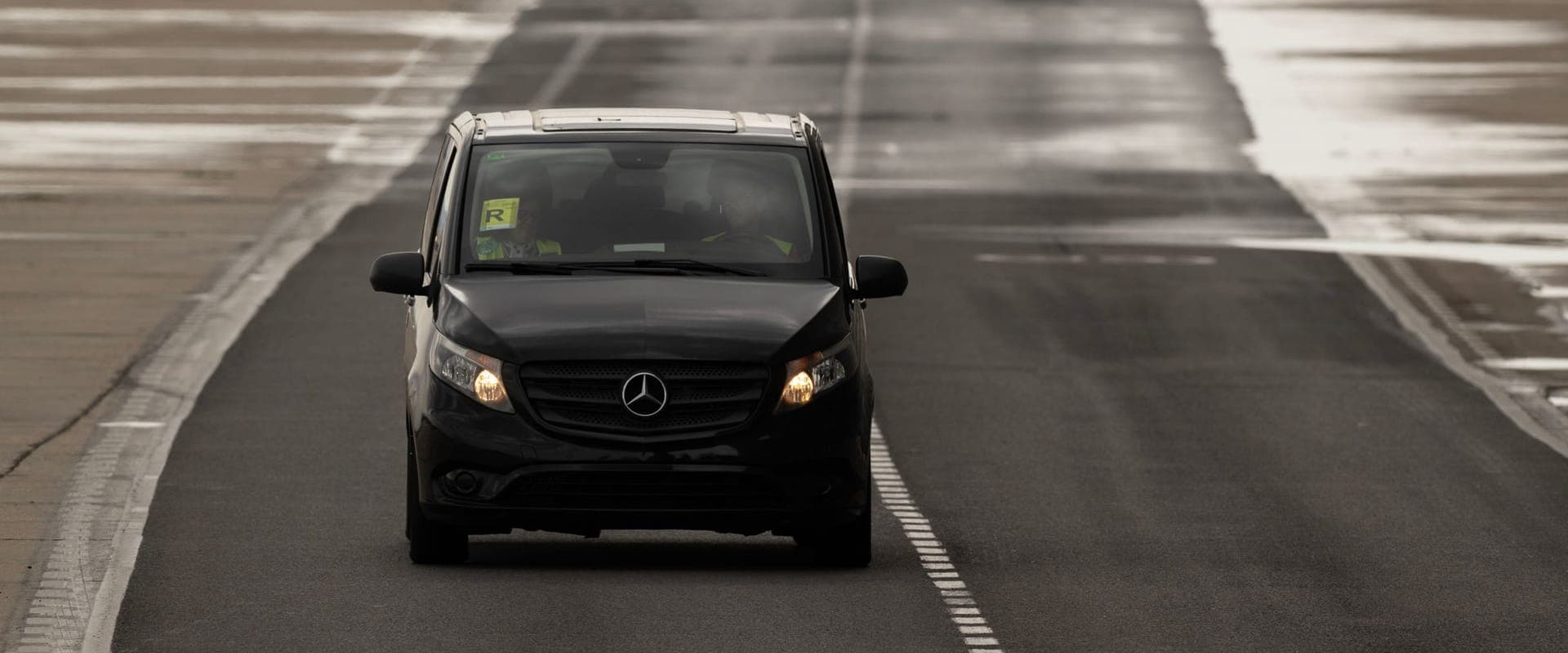 A black Mercedes Vito with its headlights on, driving across an airport apron